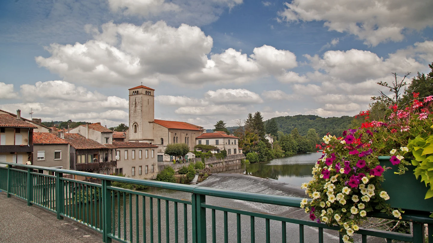À Roquecourbe, les maisons et le clocher bordent l’Agout, vus depuis un pont à rambarde verte fleurie.