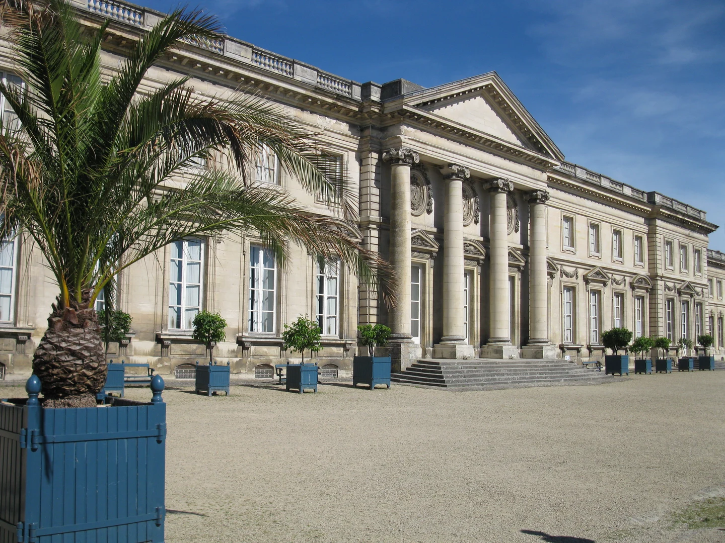Façade à colonnes du château de Compiègne, côté parc, devant une cour et des palmiers en bacs.