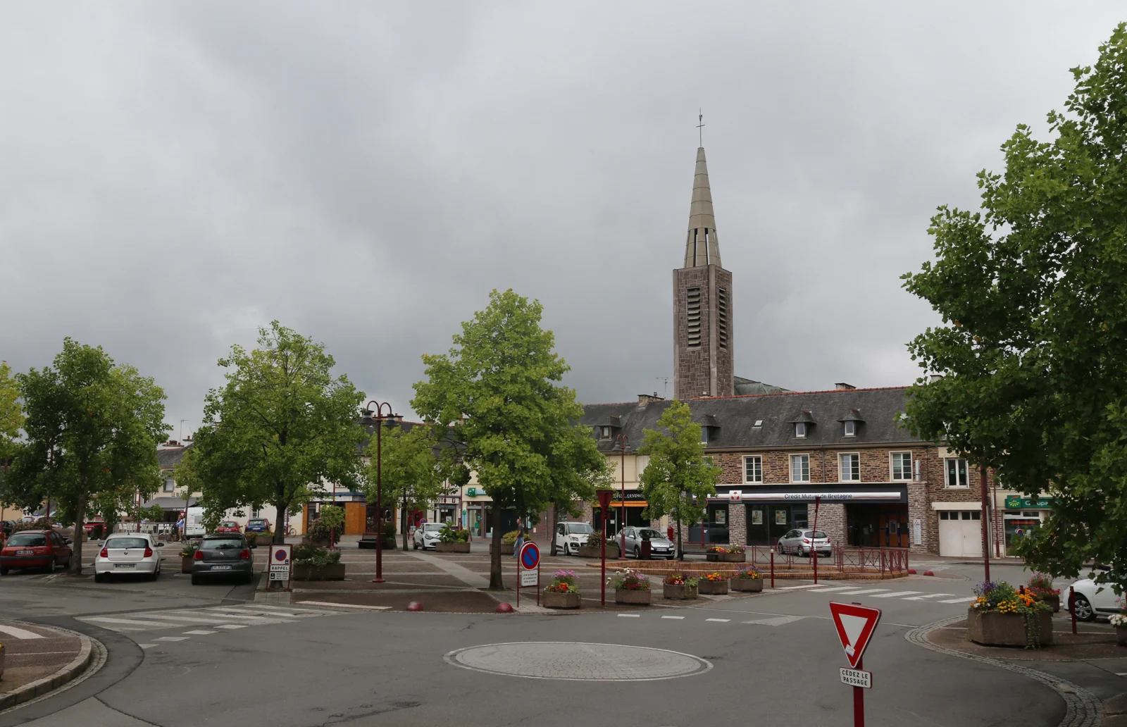 La place principale de Bruz, avec des arbres, des commerces et le clocher à l’arrière-plan.