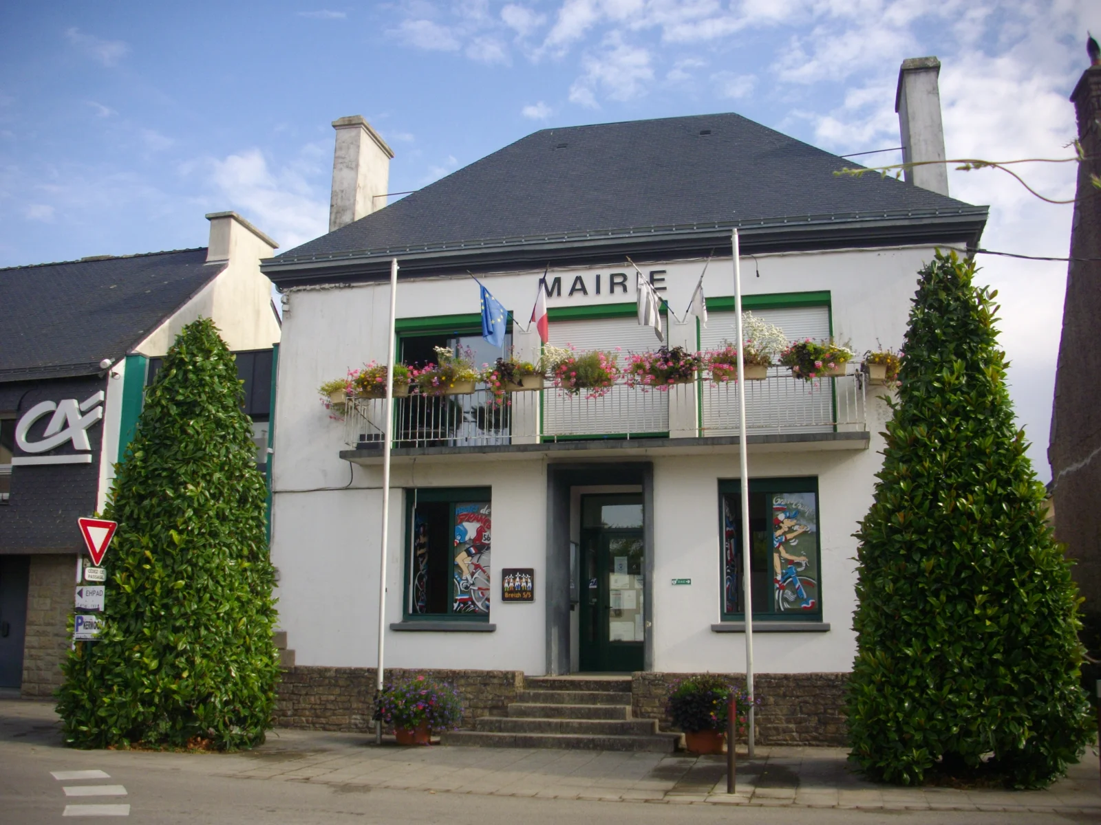 Façade blanche de la mairie de Grand-Champ, avec un balcon fleuri et des drapeaux, encadrée de deux arbres taillés.