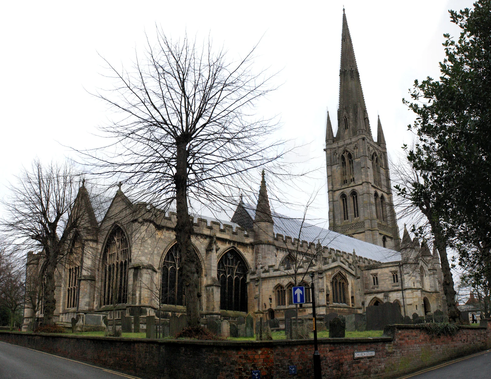 Église en pierre à haute flèche, derrière des arbres dénudés et un muret, à Grantham.