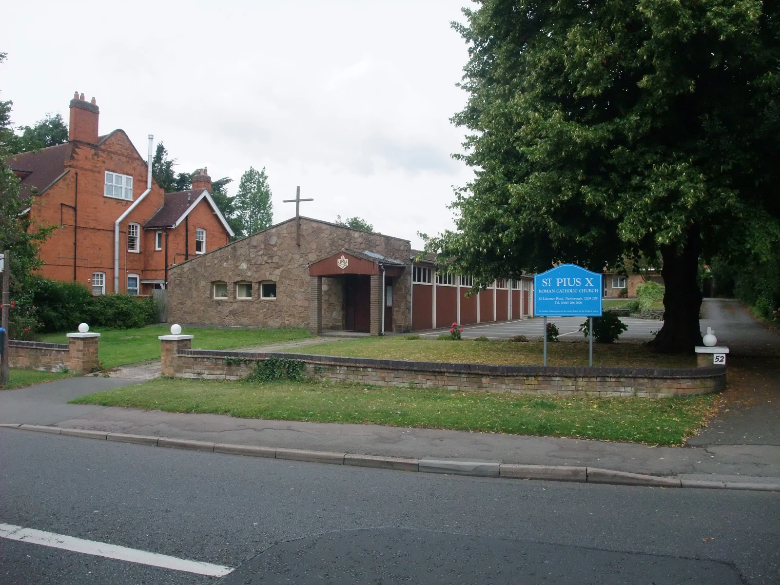 L’église basse St Pius X à Narborough, une maison en briques et de grands arbres, vus depuis la rue.