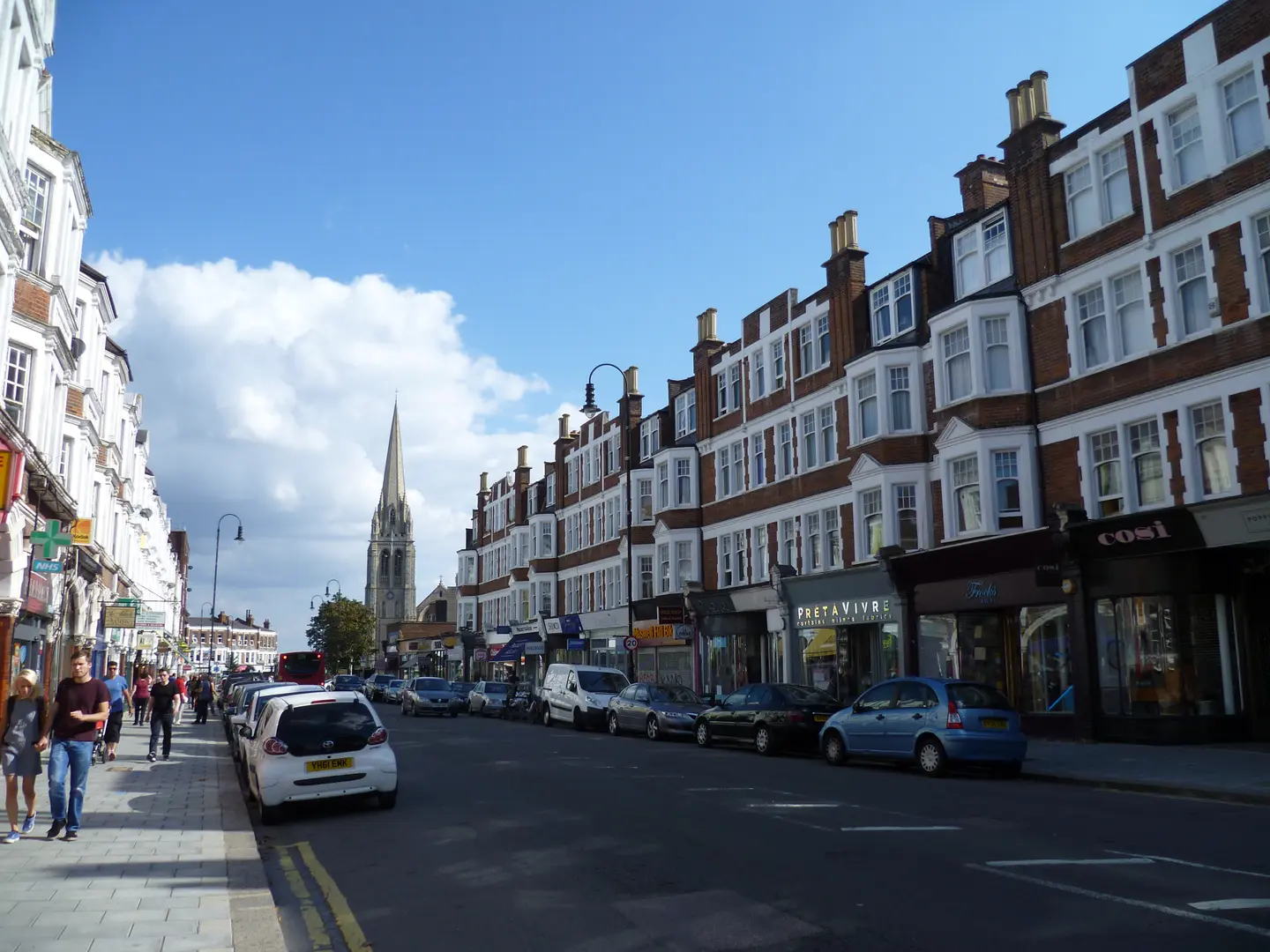 Rue commerçante de Muswell Hill, avec façades en brique et clocher à l’arrière-plan.