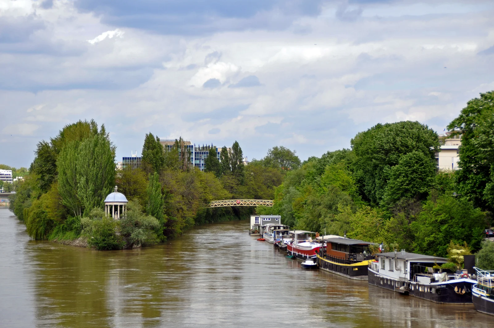 La Seine à Neuilly-sur-Seine, avec la pointe boisée de l’île de la Jatte et le temple de l’Amour à gauche, une passerelle au centre et des péniches amarrées à droite.