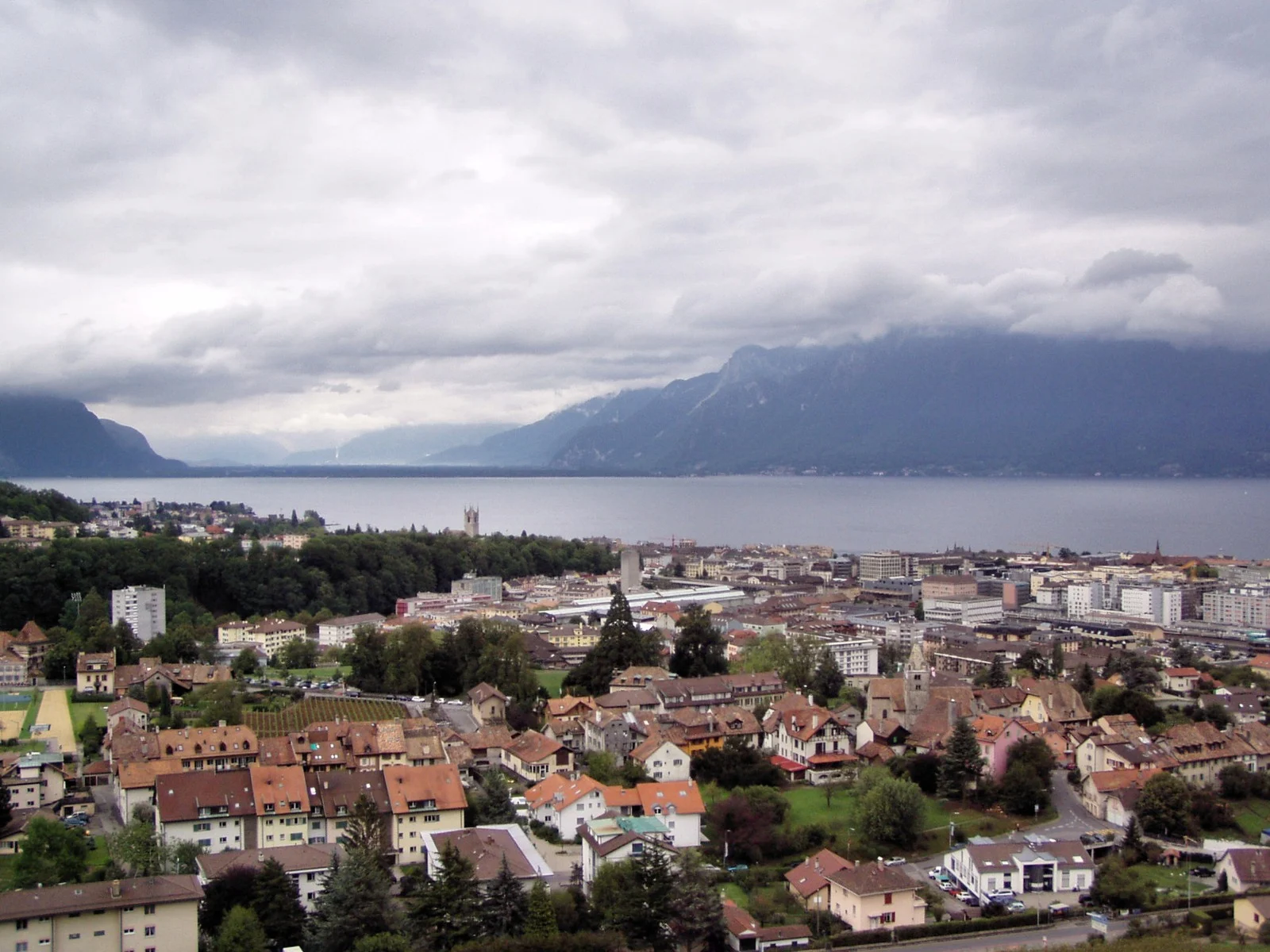 Vue générale de Corsier-sur-Vevey et de Vevey, avec le Léman et les montagnes sous un ciel couvert.