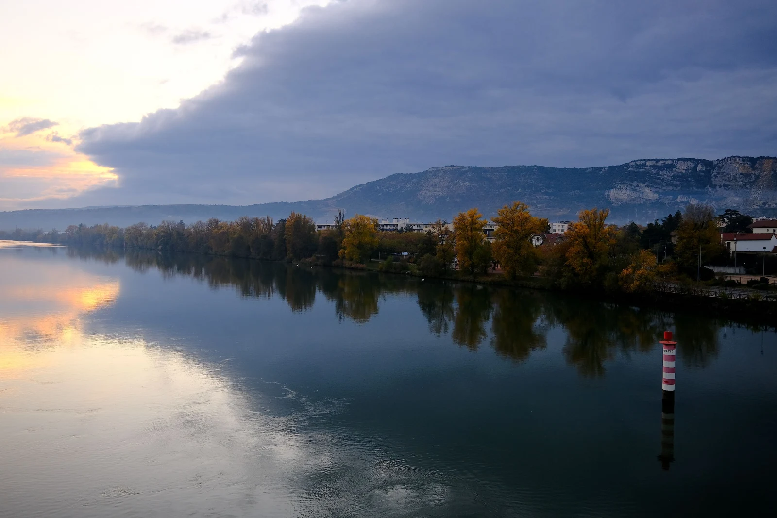 Le Rhône devant les berges arborées de Guilherand-Granges, avec des bâtiments et la montagne de Crussol au fond, à la tombée du jour.