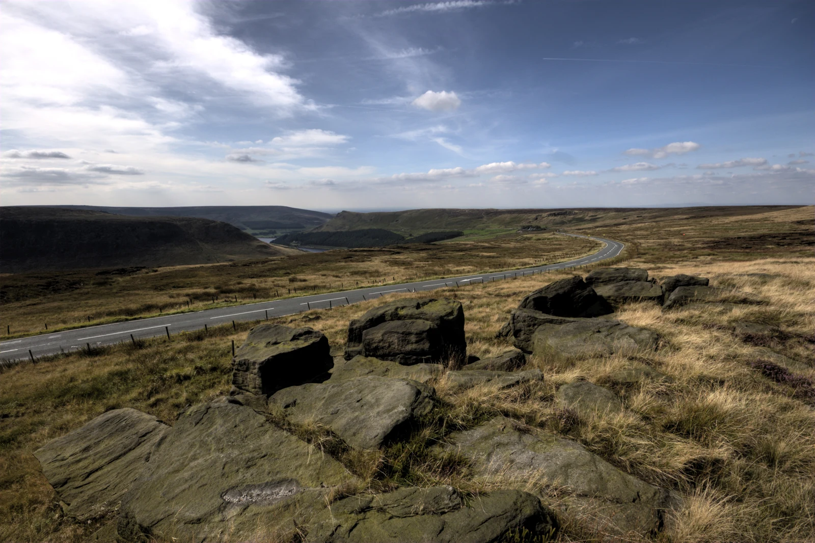 Lande de Saddleworth Moor, rochers au premier plan et route sinueuse sous un ciel bleu nuageux.