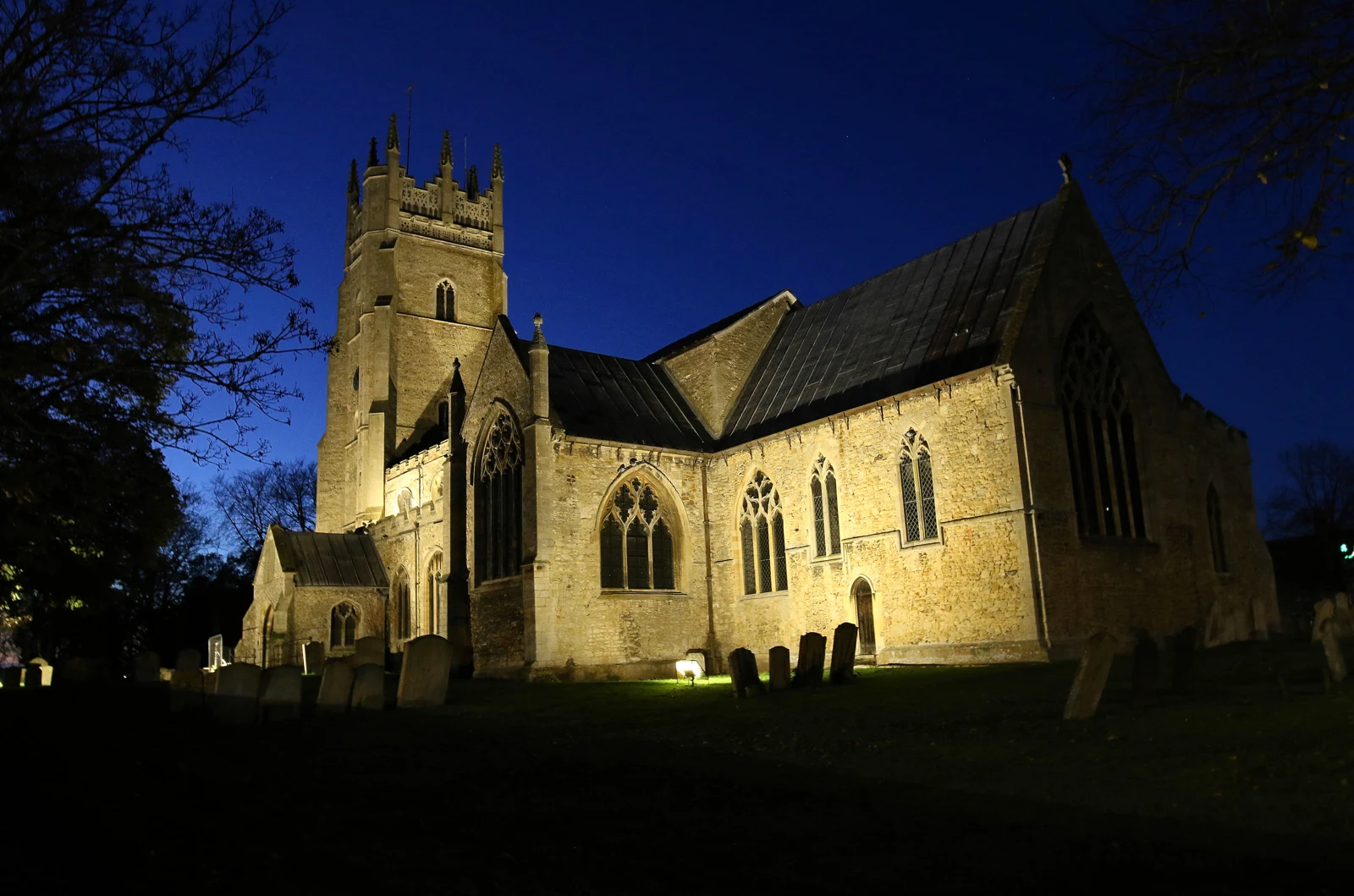 Église St Andrew de Soham éclairée la nuit, avec son clocher carré sur un ciel bleu sombre.