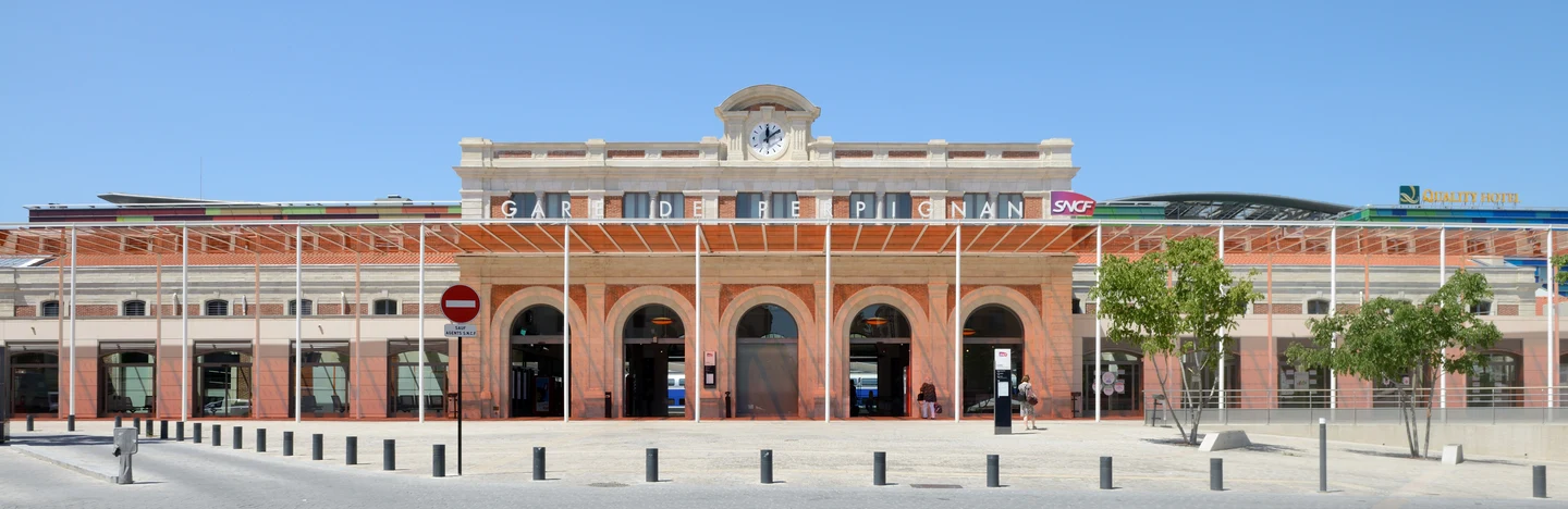 Façade de la gare de Perpignan avec son horloge centrale, ses grandes arches et son parvis ensoleillé.