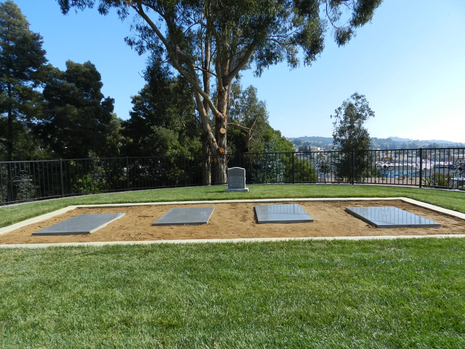 Quatre plaques commémoratives et une stèle sur une pelouse du cimetière Evergreen, avec des arbres et Oakland à l’arrière-plan.