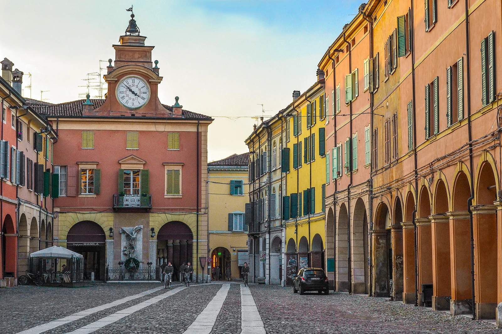 Rue pavée du corso Mazzini à Correggio, bordée de façades colorées à arcades et dominée par une horloge.