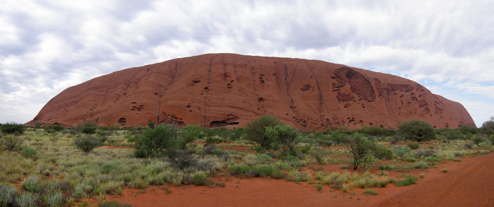 Panorama du massif rouge d’Uluru sous un ciel nuageux, avec des arbustes et un sol ocre au premier plan, sans personnes visibles.
