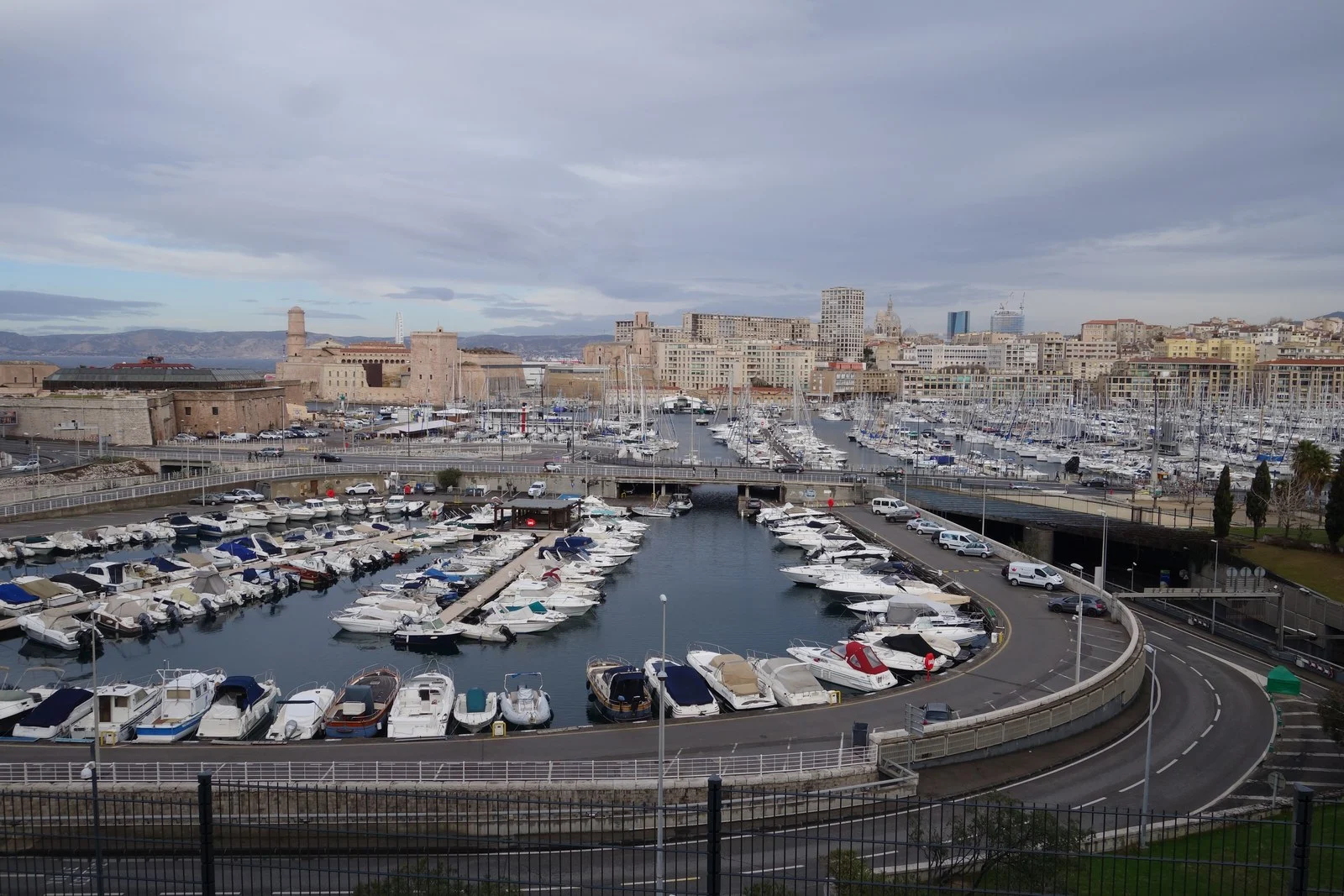 Vue en hauteur du Vieux-Port de Marseille, avec les bateaux amarrés, le fort Saint-Jean et les immeubles sous un ciel couvert.