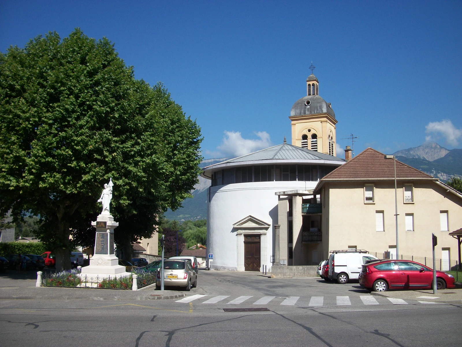La place de l’église Saint-Antoine à Villard-Bonnot, avec le monument aux morts, les arbres et les montagnes à l’arrière-plan.