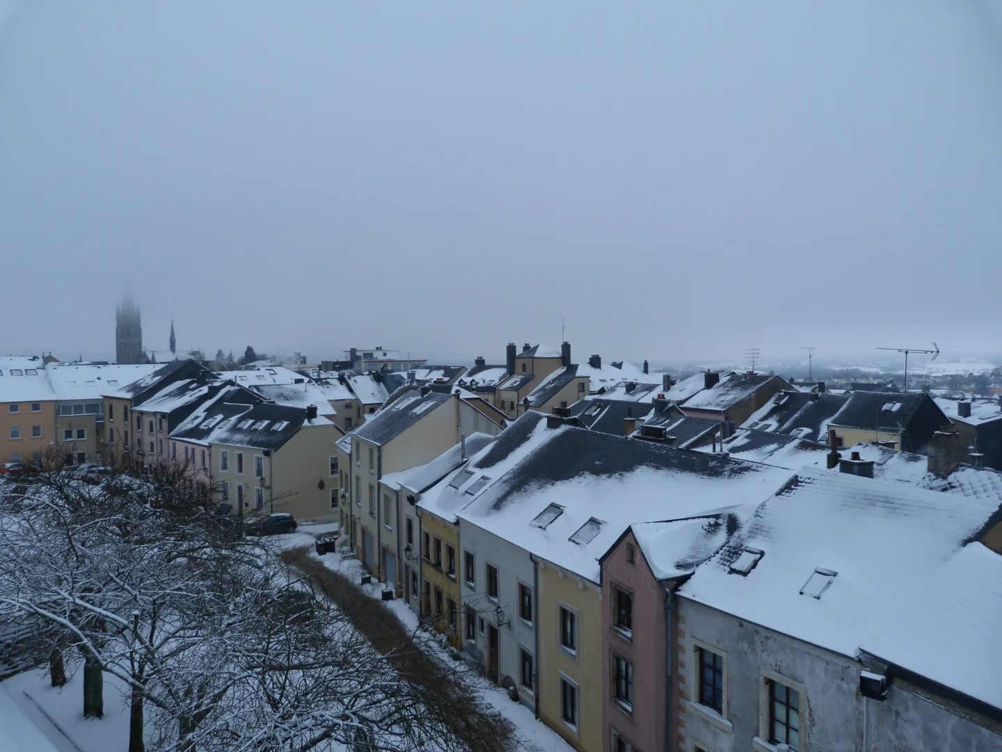 Toits enneigés d’Arlon, avec le clocher de l’église Saint-Martin dans le brouillard au loin.