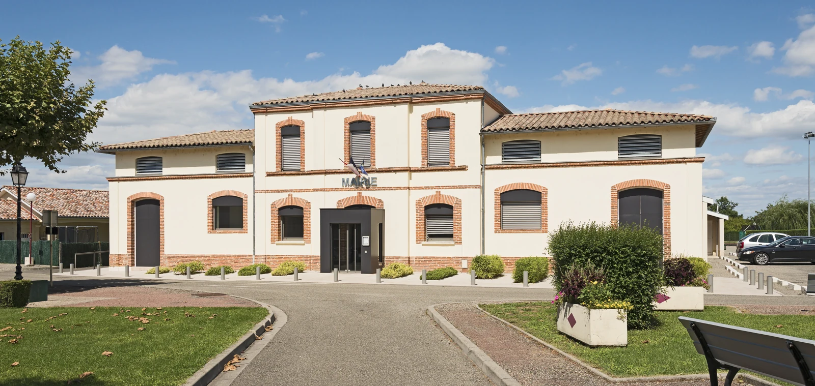 Façade de la mairie de Montbartier, bâtiment clair aux encadrements de briques, précédé de pelouses et de massifs fleuris.