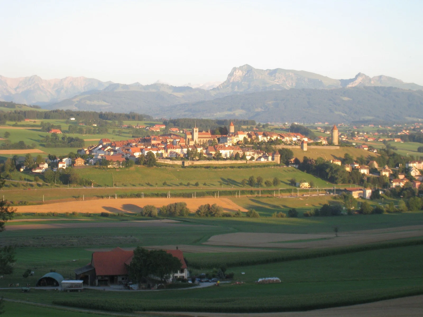 Vue générale de Romont et de ses remparts sur une colline, entourée de champs, avec les montagnes à l’arrière-plan.