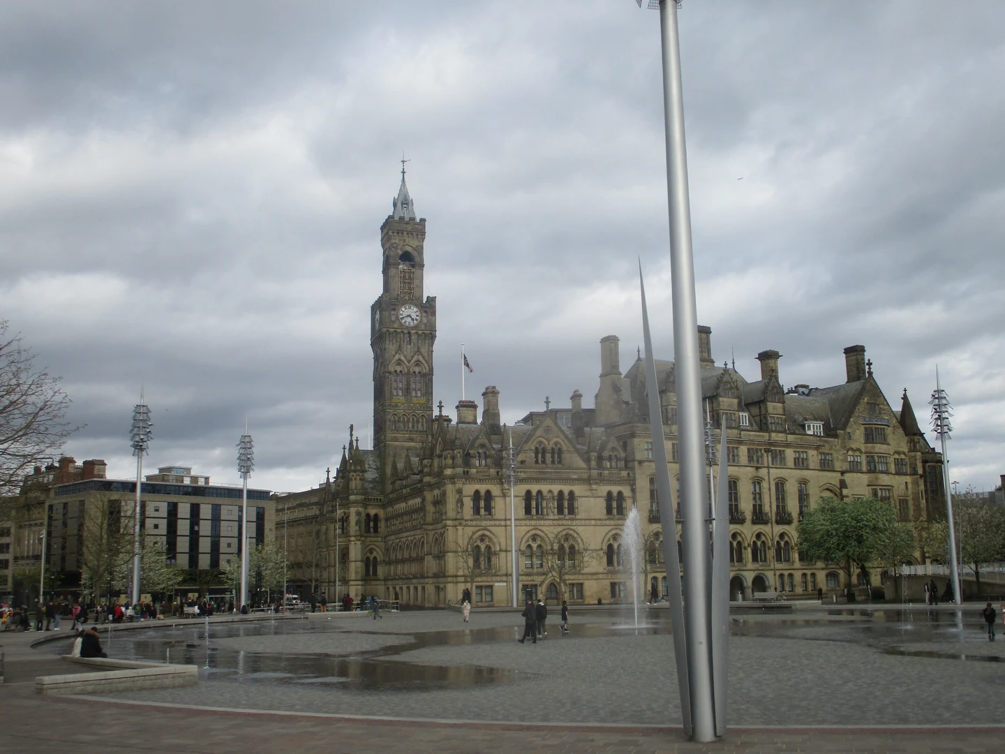 L’hôtel de ville de Bradford et sa tour de l’horloge, devant une place avec des fontaines, sous un ciel couvert.