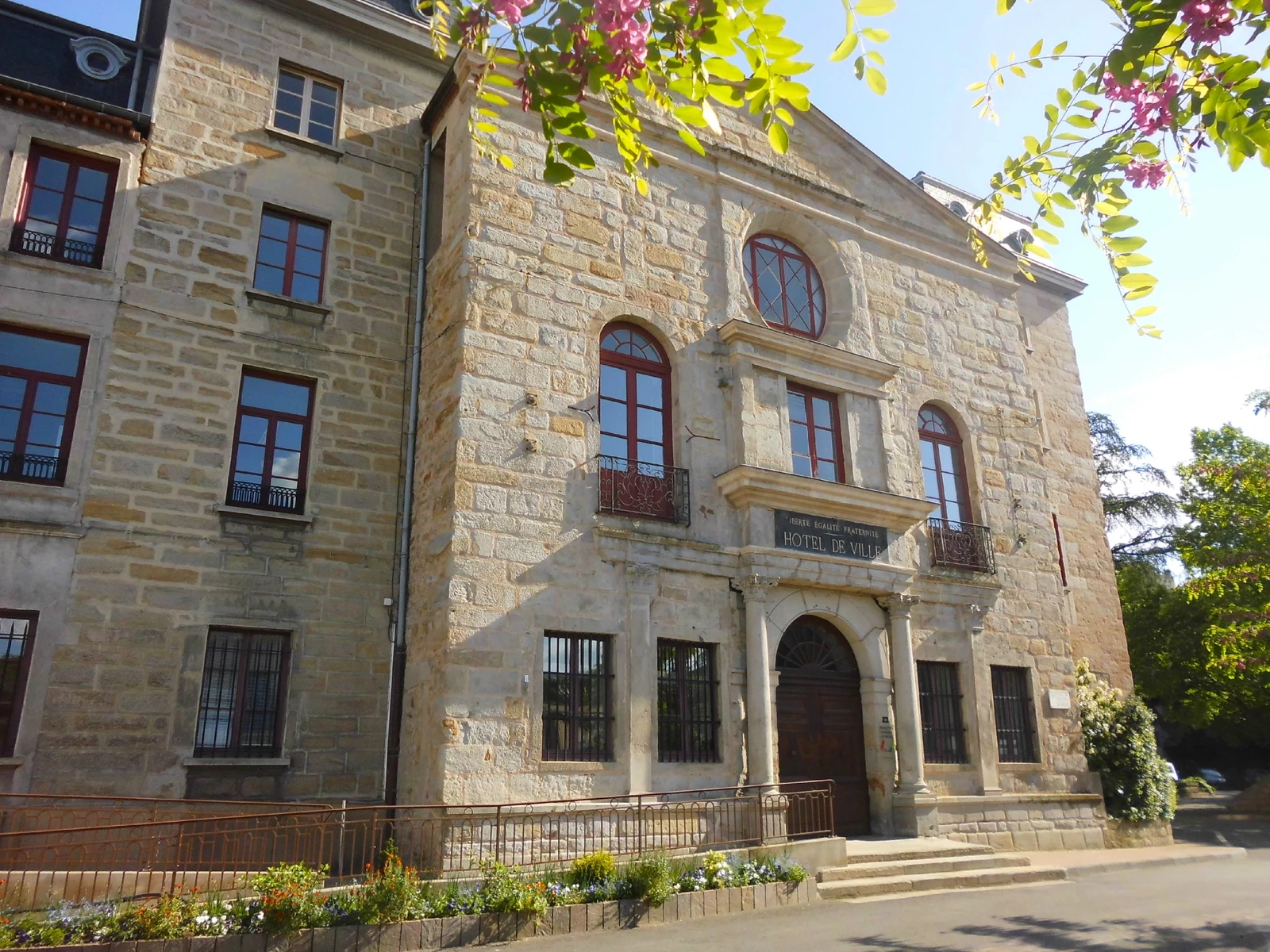 La façade de pierre de la mairie de Saint-Chamond, avec son entrée centrale, ses fenêtres rouges et des branches fleuries au premier plan.