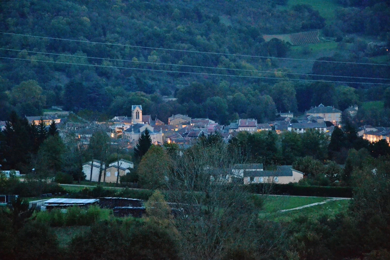 Vue de Lagnieu à la tombée du jour, avec les maisons et le clocher au pied d’une colline boisée.